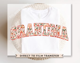 May include: White t-shirt with a floral print design that says "Grandma" in a bold, orange font. The text is outlined in a darker orange. The shirt is on a white wicker surface. Direct to film transfer.