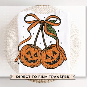 May include: White t-shirt featuring a Halloween-themed design. The design includes two orange pumpkin faces with black cut-out eyes and mouths, connected by brown stems and adorned with orange ribbons and green leaves. The text "DIRECT TO FILM TRANSFER" is at the bottom.
