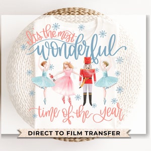 May include: A white t-shirt with a pink and blue watercolor design featuring a nutcracker and ballerinas. The text "It's the most wonderful time of the year" is written in pink and blue cursive lettering. The words "Direct to Film Transfer" are printed in black at the bottom of the shirt.