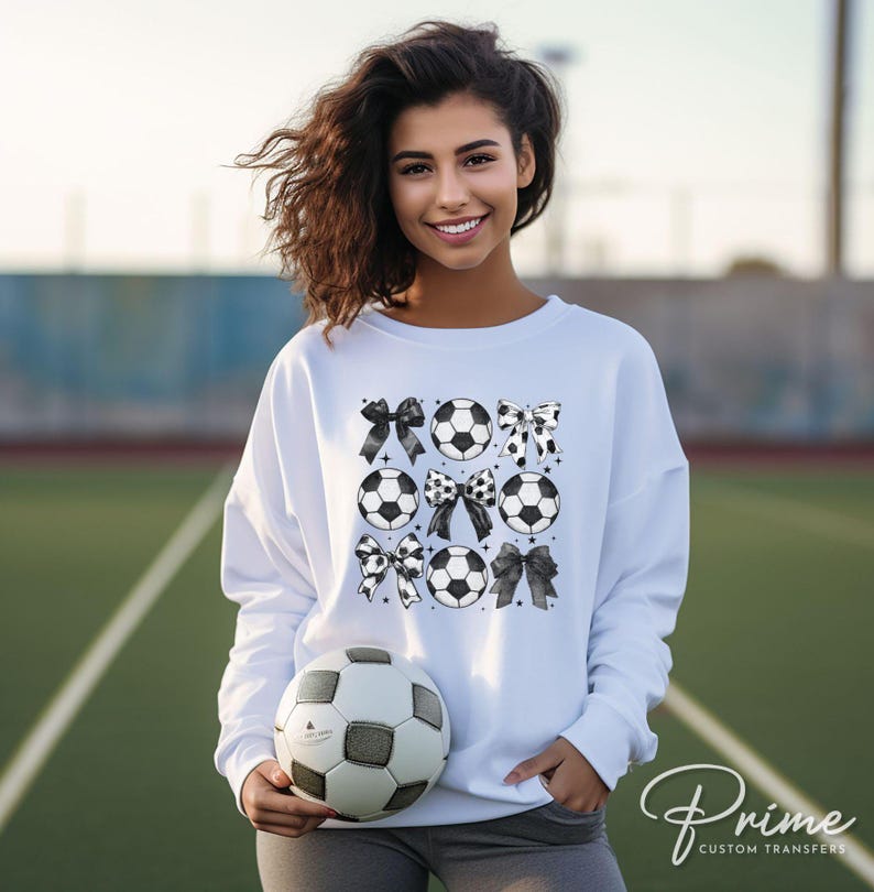 Puede incluir: Sudadera blanca con un dise&ntilde;o de bal&oacute;n de f&uacute;tbol y lazos. El gr&aacute;fico incluye balones de f&uacute;tbol y lazos en blanco y negro. Se sostiene un bal&oacute;n de f&uacute;tbol en las manos. El fondo es un campo verde.