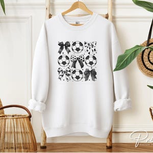 Puede incluir: Sudadera blanca con un dise&ntilde;o de bal&oacute;n de f&uacute;tbol y lazos en blanco y negro. El gr&aacute;fico presenta balones de f&uacute;tbol y lazos en un patr&oacute;n de cuadr&iacute;cula. La sudadera est&aacute; colgada en una percha de madera.