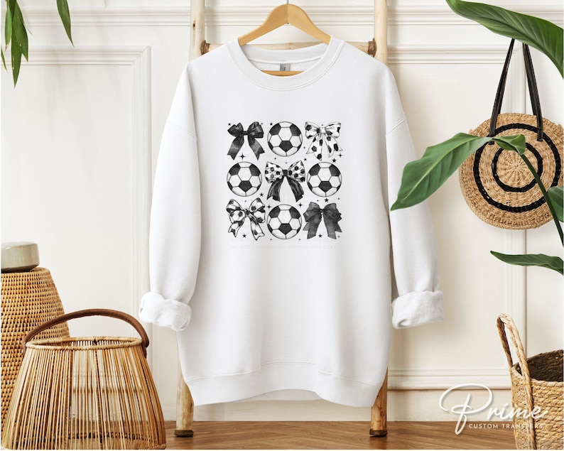 Puede incluir: Sudadera blanca con un dise&ntilde;o de bal&oacute;n de f&uacute;tbol y lazos en blanco y negro. El gr&aacute;fico presenta balones de f&uacute;tbol y lazos en un patr&oacute;n de cuadr&iacute;cula. La sudadera est&aacute; colgada en una percha de madera.