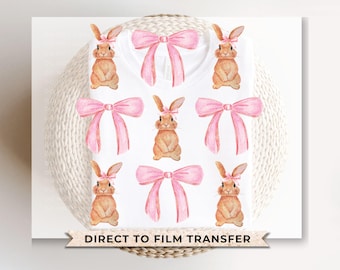 Può includere: Maglietta bianca con un fiocco rosa e una stampa di coniglio marrone. La stampa si ripete su tutta la maglietta. "Direct to Film Transfer" è stampato sul fondo della maglietta.