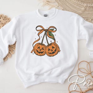 May include: White sweatshirt featuring a Halloween-themed graphic. The design includes two orange jack-o'-lanterns with smiling faces, connected by stems and a bow. The graphic is in a vintage style.