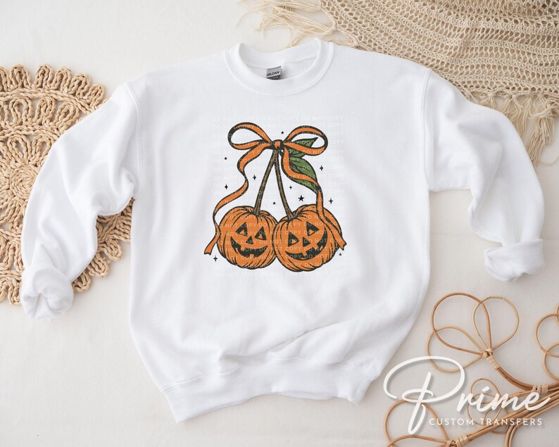 May include: White sweatshirt featuring a Halloween-themed graphic. The design includes two orange jack-o'-lanterns with smiling faces, connected by stems and a bow. The graphic is in a vintage style.