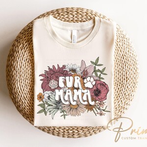May include: Cream-colored t-shirt with the text "FUR MAMA" in a retro font, surrounded by a floral design with a paw print. The flowers include shades of pink, orange, and white. The shirt is displayed on a woven, circular surface.
