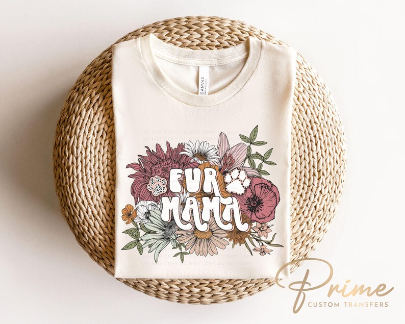 May include: Cream-colored t-shirt with the text "FUR MAMA" in a retro font, surrounded by a floral design with a paw print. The flowers include shades of pink, orange, and white. The shirt is displayed on a woven, circular surface.