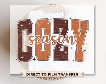 May include: A white t-shirt with a brown and orange graphic that says "Cozy season" with stars around the text. The text is in a vintage style font. The shirt is a direct to film transfer.