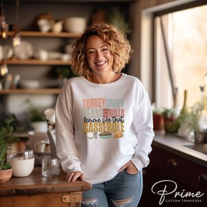 May include: White sweatshirt with a Thanksgiving themed graphic that says "Turkey Gravy Beans Rolls lemme see that Casserole". The graphic is in a retro style with different colors and fonts.
