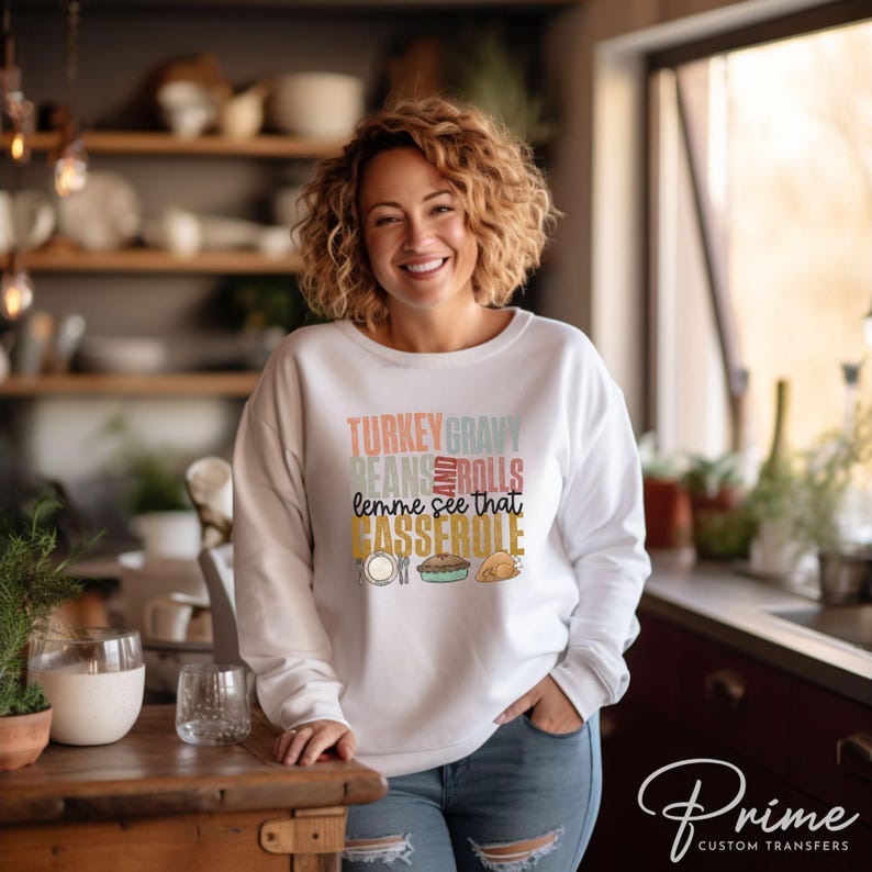 May include: White sweatshirt with a Thanksgiving themed graphic that says "Turkey Gravy Beans Rolls lemme see that Casserole". The graphic is in a retro style with different colors and fonts.