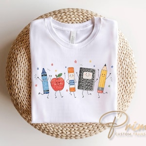 May include: A white t-shirt featuring a colorful graphic print of five anthropomorphic school supplies including a blue crayon, a red apple, an orange glue stick, a black composition notebook, and a yellow pencil, all with smiling faces.