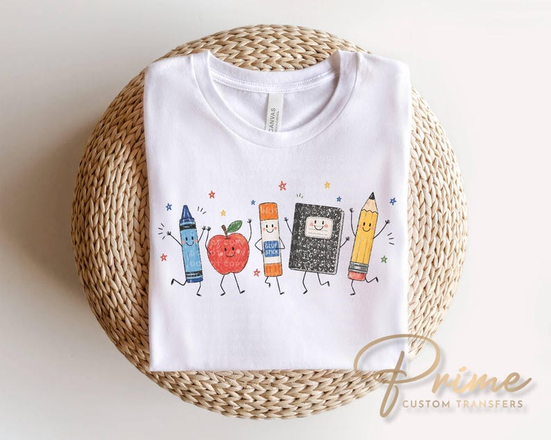 May include: A white t-shirt featuring a colorful graphic print of five anthropomorphic school supplies including a blue crayon, a red apple, an orange glue stick, a black composition notebook, and a yellow pencil, all with smiling faces.