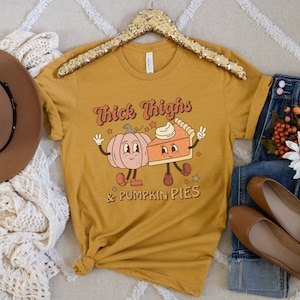 K&ouml;nnte beinhalten: Ein senfgelbes T-Shirt mit einem Aufdruck einer K&uuml;rbis und einem St&uuml;ck K&uuml;rbiskuchen mit dem Text "Thick Thighs & Pumpkin Pies".