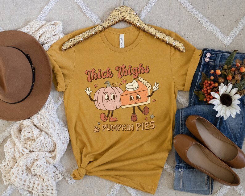 K&ouml;nnte beinhalten: Ein senfgelbes T-Shirt mit einem Aufdruck einer K&uuml;rbis und einem St&uuml;ck K&uuml;rbiskuchen mit dem Text "Thick Thighs & Pumpkin Pies".