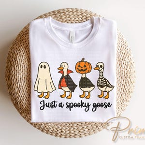 May include: White t-shirt featuring a Halloween-themed graphic of four cartoon geese dressed as a ghost, vampire, jack-o'-lantern, and skeleton. The text "Just a spooky goose" is printed below the geese.