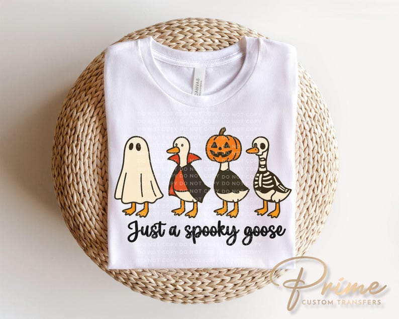 May include: White t-shirt featuring a Halloween-themed graphic of four cartoon geese dressed as a ghost, vampire, jack-o'-lantern, and skeleton. The text "Just a spooky goose" is printed below the geese.