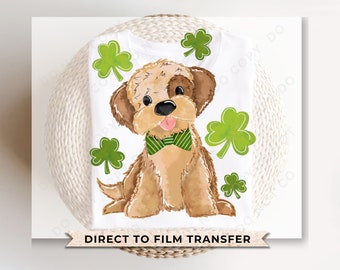 Peut inclure: Un t-shirt blanc avec un chien brun et blanc portant un nœud papillon vert et blanc. Le chien est entouré de feuilles de trèfle vertes. Le texte "DIRECT TO FILM TRANSFER" est imprimé en bas du t-shirt.
