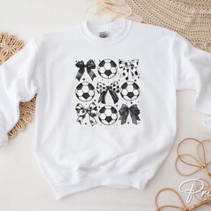 Puede incluir: Sudadera blanca con un dise&ntilde;o en blanco y negro de balones de f&uacute;tbol y lazos. Los lazos tienen varios patrones, incluyendo lunares y colores lisos. La sudadera es de cuello redondo.