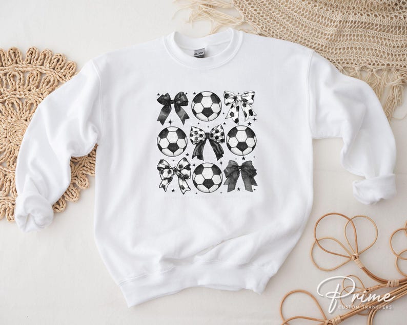 Puede incluir: Sudadera blanca con un dise&ntilde;o en blanco y negro de balones de f&uacute;tbol y lazos. Los lazos tienen varios patrones, incluyendo lunares y colores lisos. La sudadera es de cuello redondo.