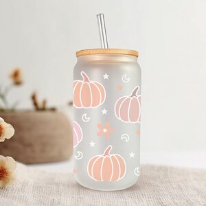 K&ouml;nnte beinhalten: Ein gefrosteter Glasbecher mit einem Holzdeckel und einem Strohhalm. Der Becher hat ein Muster aus rosa und orangefarbenen K&uuml;rbissen, Sternen und Monden.