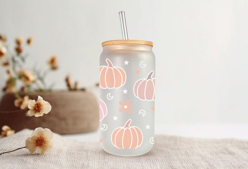 K&ouml;nnte beinhalten: Ein gefrosteter Glasbecher mit einem Holzdeckel und einem Strohhalm. Der Becher hat ein Muster aus rosa und orangefarbenen K&uuml;rbissen, Sternen und Monden.
