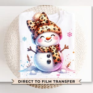 May include: A white t-shirt with a cartoon snowman wearing a leopard print hat and scarf. The text "DIRECT TO FILM TRANSFER" is printed at the bottom of the shirt.