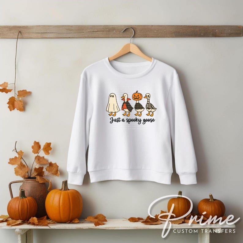 May include: White sweatshirt with a Halloween-themed graphic of four geese dressed as a ghost, vampire, pumpkin, and skeleton. The text "Just a spooky goose" is printed below the geese. The sweatshirt is hanging on a wooden hanger.