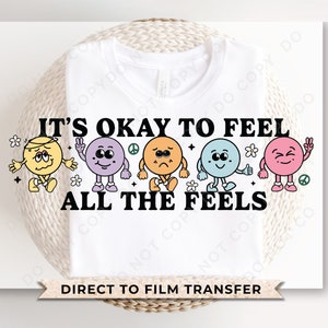 May include: White t-shirt with the text "It's okay to feel all the feels" and five colorful cartoon faces with different expressions. The text "Direct to film transfer" is at the bottom of the shirt.