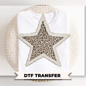 May include: White t-shirt with a large star design. The star features a leopard print center and a frayed, light-colored border. The text "DTF TRANSFER" is displayed on a banner below the shirt.