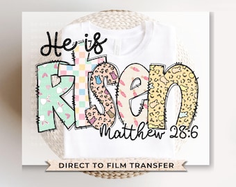 May include: White t-shirt with a colorful "He is Risen" design with Easter-themed patterns. The text "Matthew 28:6" is below the design. The shirt is a direct to film transfer.