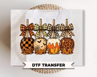 May include: A white t-shirt featuring a DTF transfer graphic of four caramel apples on sticks, each topped with a patterned bow. The apples display orange and black buffalo plaid, a bat and spiderweb print, candy corn colours, and leopard print.