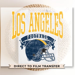 May include: A white t-shirt with a distressed yellow and blue graphic design that says "Los Angeles Football" with a football helmet graphic. The text "Direct to Film Transfer" is printed on a yellow banner at the bottom of the shirt.