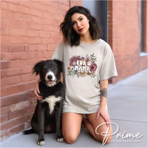 May include: A light beige t-shirt with the text "FUR MAMA" in a floral design, featuring a paw print. A black and white dog sits next to the person wearing the shirt.