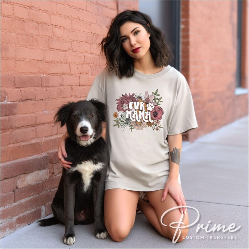 May include: A light beige t-shirt with the text "FUR MAMA" in a floral design, featuring a paw print. A black and white dog sits next to the person wearing the shirt.