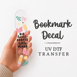 May include: A clear, rectangular bookmark decal featuring the text BIG heart SHAPES little MINDS in black lettering, surrounded by colorful illustrations of yellow pencils, pink books, green leaves, and pink bows, with a hole at the top.