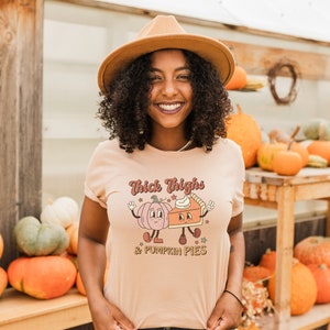 K&ouml;nnte beinhalten: Eine Frau tr&auml;gt einen hellbraunen Hut und ein pfirsichfarbenes T-Shirt mit dem Text "Thick Thighs & Pumpkin Pies" und einem Cartoon-Bild von einem K&uuml;rbis und einer Torte.
