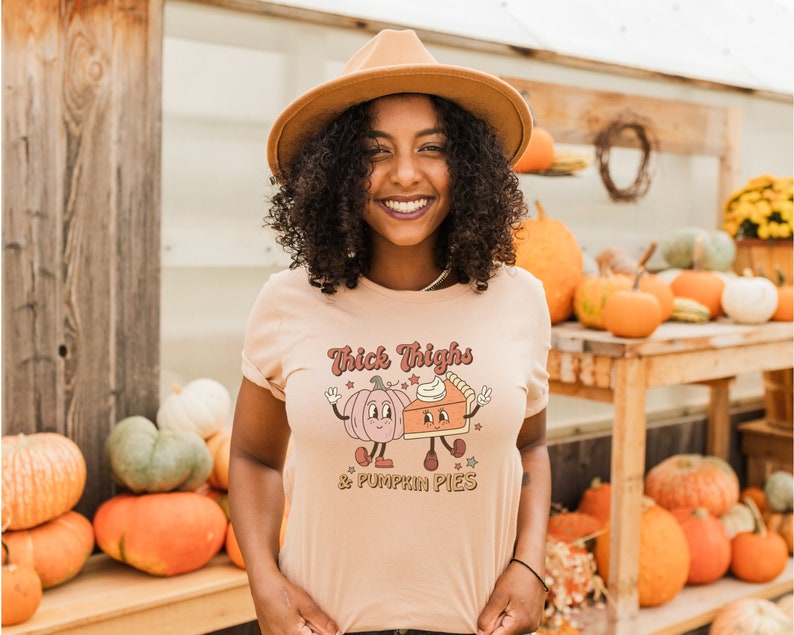 K&ouml;nnte beinhalten: Eine Frau tr&auml;gt einen hellbraunen Hut und ein pfirsichfarbenes T-Shirt mit dem Text "Thick Thighs & Pumpkin Pies" und einem Cartoon-Bild von einem K&uuml;rbis und einer Torte.