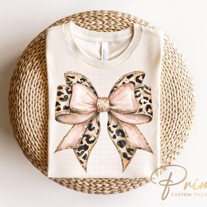 May include: A white t-shirt with a large pink and brown leopard print bow with gold glitter accents. The bow is centered on the shirt.