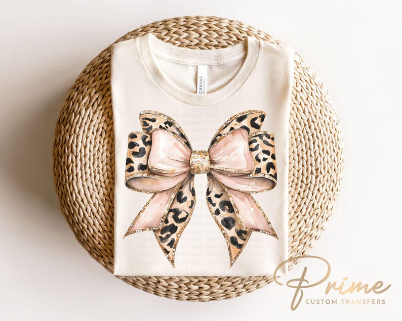 May include: A white t-shirt with a large pink and brown leopard print bow with gold glitter accents. The bow is centered on the shirt.