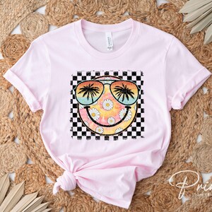 May include: Pink t-shirt with a graphic of a smiley face wearing sunglasses with palm trees reflected in the lenses. The smiley face is surrounded by a checkered pattern and daisies.