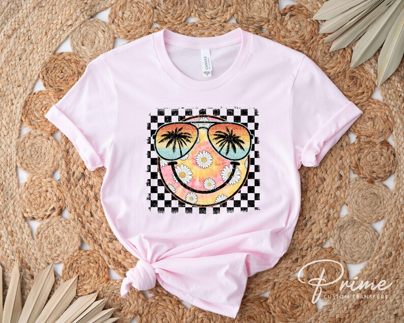 May include: Pink t-shirt with a graphic of a smiley face wearing sunglasses with palm trees reflected in the lenses. The smiley face is surrounded by a checkered pattern and daisies.