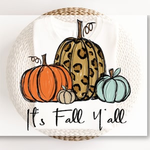 May include: A white t-shirt with a graphic of three pumpkins. The largest pumpkin is brown and black with a leopard print. The middle pumpkin is orange and the smallest pumpkin is light brown. The text "It's Fall Y'all" is written below the pumpkins.