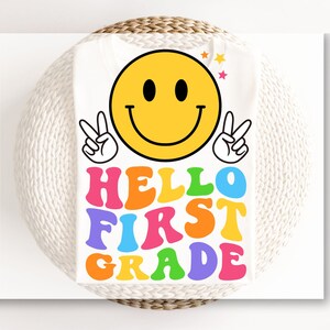 May include: A white t-shirt featuring a large yellow smiley face with two hands making peace signs. Below the smiley face, the text HELLO FIRST GRADE is printed in a bold, wavy, multi-colored font including pink, orange, blue, green, and yellow.