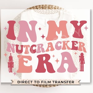May include: Pink and red graphic design on a white t-shirt that says "In My Nutcracker Era" with a silhouette of a nutcracker on each side. The text is in a retro font style. The t-shirt is for direct to film transfer.