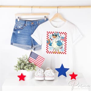 K&ouml;nnte beinhalten: Wei&szlig;es T-Shirt mit einem rot-wei&szlig;-blauen karierten Hintergrund, auf dem eine Cartoon-Gans mit Sonnenbrille und einem rot-wei&szlig;-blauen Hut zu sehen ist, die einen Hotdog h&auml;lt. Die Gans steht auf einem rot-wei&szlig;-blauen karierten Hintergrund mit Feuerwerk und einer kleinen amerikanischen Flagge.