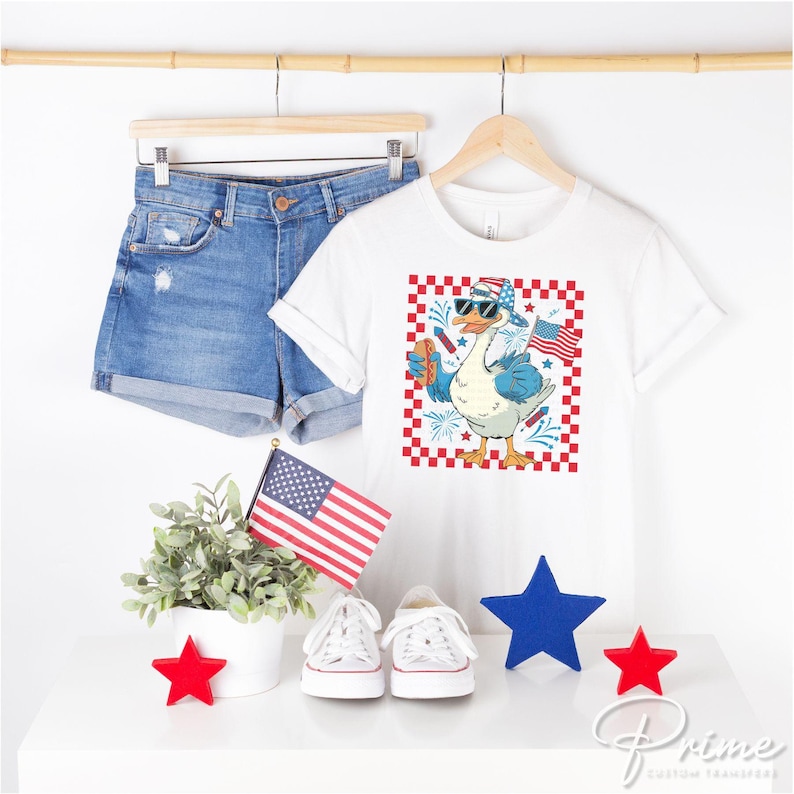 K&ouml;nnte beinhalten: Wei&szlig;es T-Shirt mit einem rot-wei&szlig;-blauen karierten Hintergrund, auf dem eine Cartoon-Gans mit Sonnenbrille und einem rot-wei&szlig;-blauen Hut zu sehen ist, die einen Hotdog h&auml;lt. Die Gans steht auf einem rot-wei&szlig;-blauen karierten Hintergrund mit Feuerwerk und einer kleinen amerikanischen Flagge.