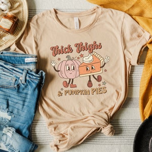 K&ouml;nnte beinhalten: Ein beiges T-Shirt mit einem Aufdruck einer K&uuml;rbis und einem St&uuml;ck K&uuml;rbiskuchen mit dem Text "Thick Thighs & Pumpkin Pies".