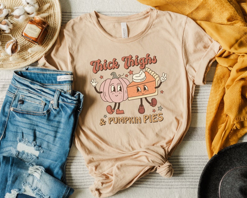 K&ouml;nnte beinhalten: Ein beiges T-Shirt mit einem Aufdruck einer K&uuml;rbis und einem St&uuml;ck K&uuml;rbiskuchen mit dem Text "Thick Thighs & Pumpkin Pies".