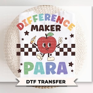 May include: A white t-shirt featuring a colorful graphic of a red apple character holding a pencil and making a peace sign. The text reads DIFFERENCE MAKER PARA. The design includes a black and white checkered stripe and small dark brown stars.
