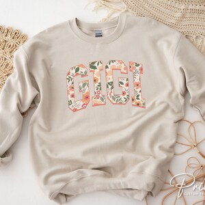 May include: A beige sweatshirt with the word "GIGI" printed in large, bold letters. The letters are filled with a floral pattern in shades of pink, orange, and green.